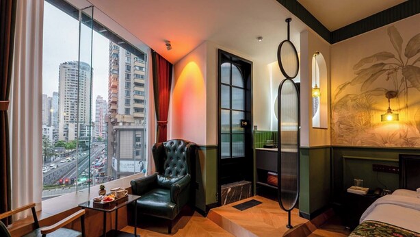 A hotel room with a large window, a black leather armchair, a small table, a bed, and a wall-mounted light fixture. - Xigaodi Art Homestay Hotel  (Guiyang)