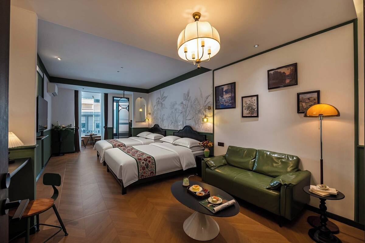 A hotel room with a bed, a green leather sofa, a dining table with a tray of food, a lamp, and a chandelier.