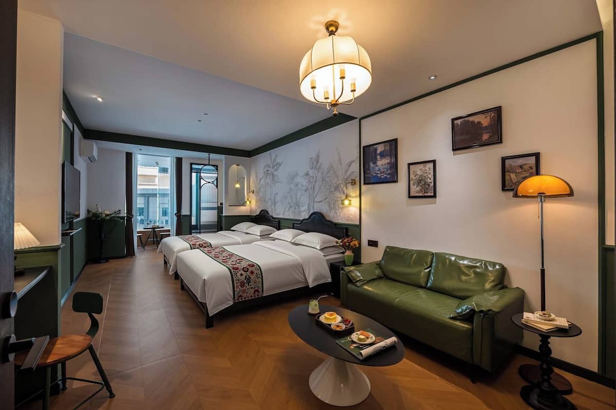 A hotel room with a bed, a green leather sofa, a dining table with a tray of food, a lamp, and a chandelier.