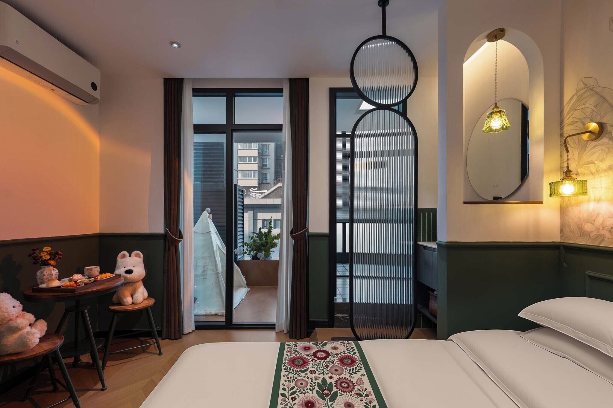 A hotel room with a bed, a small table with a teddy bear, a chair, a balcony with a view of buildings, and a decorative wall with a mirror and light fixture.