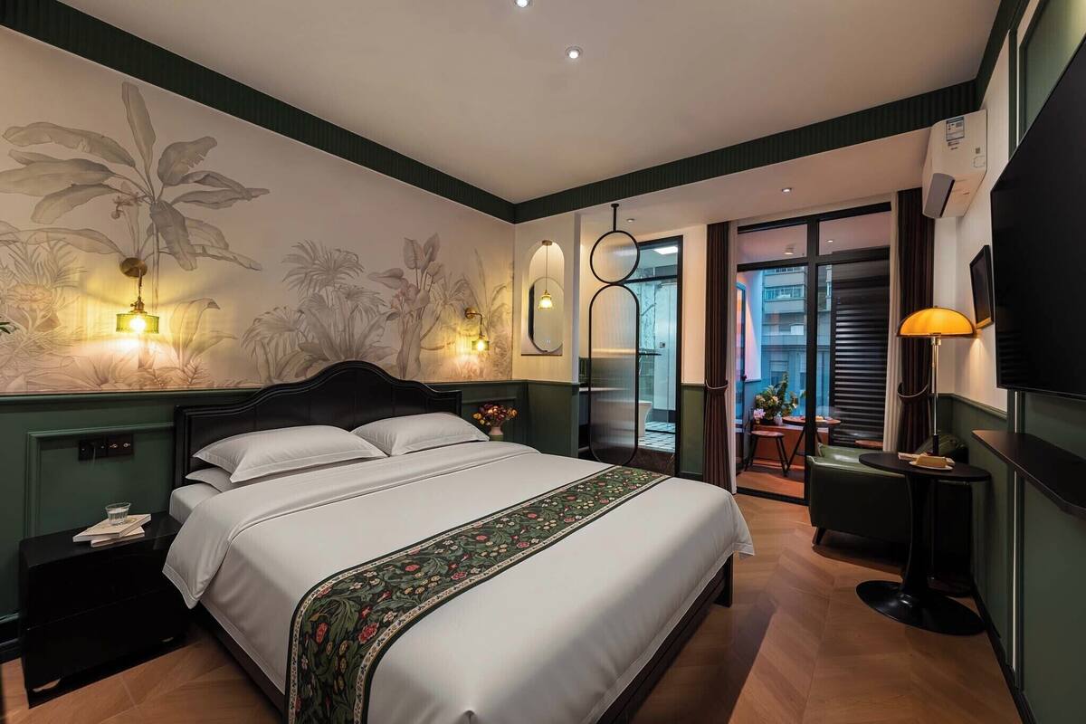 A hotel room with a large bed, bedside tables, a wall-mounted TV, a small table with a lamp, and a view through a glass door to an outdoor area.