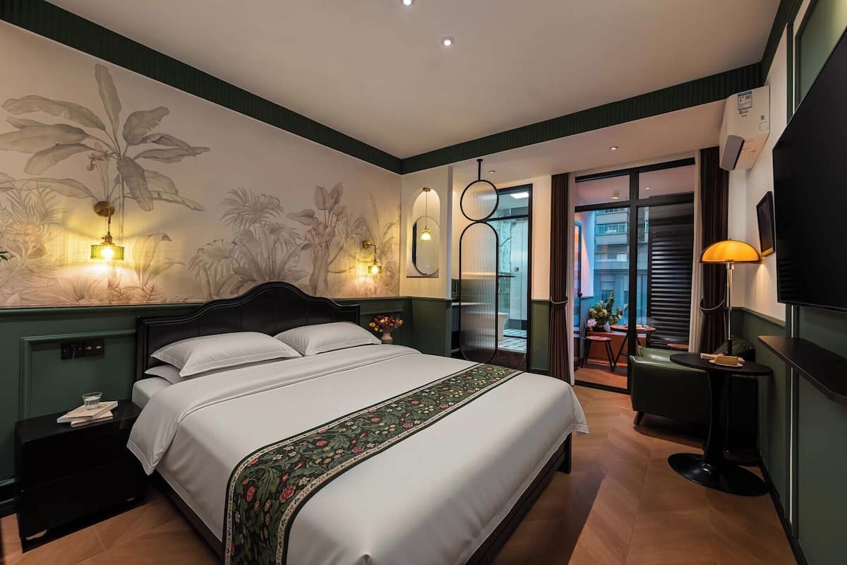 A hotel room with a large bed, bedside tables, a wall-mounted TV, a small table with a lamp, and a view through a glass door to an outdoor area.