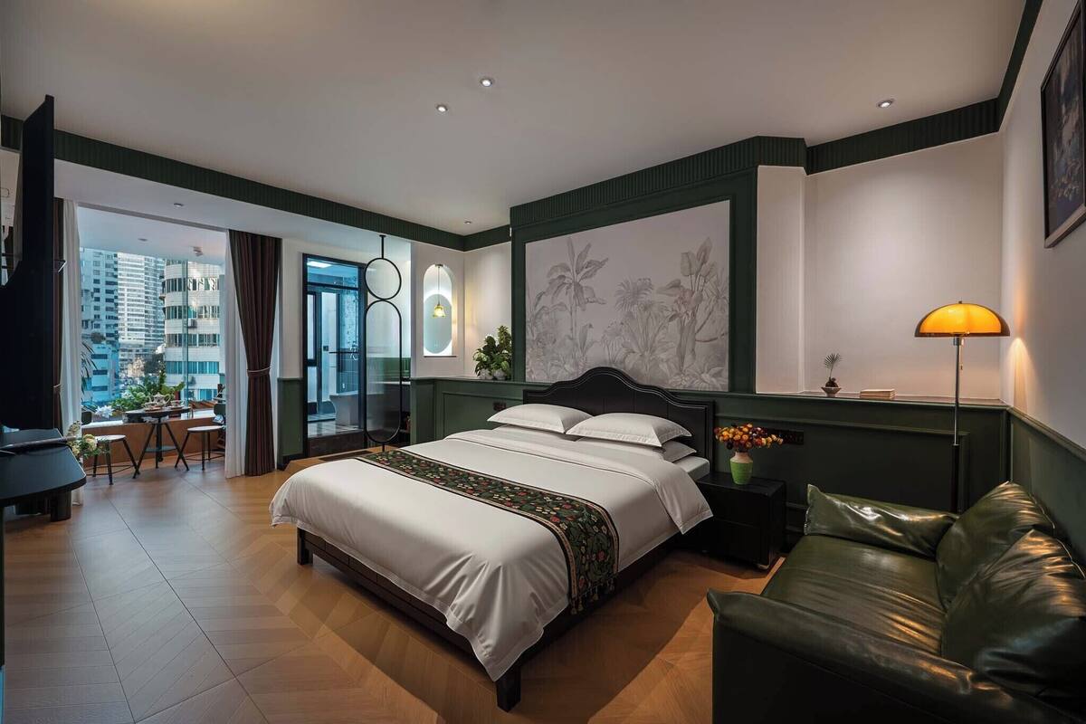 A modern hotel room with a large bed, a green leather sofa, a desk with a chair, and a view of the city.