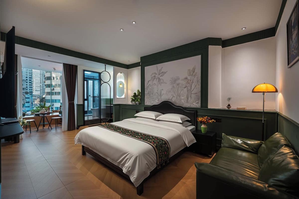 A modern hotel room with a large bed, a green leather sofa, a desk with a chair, and a view of the city.