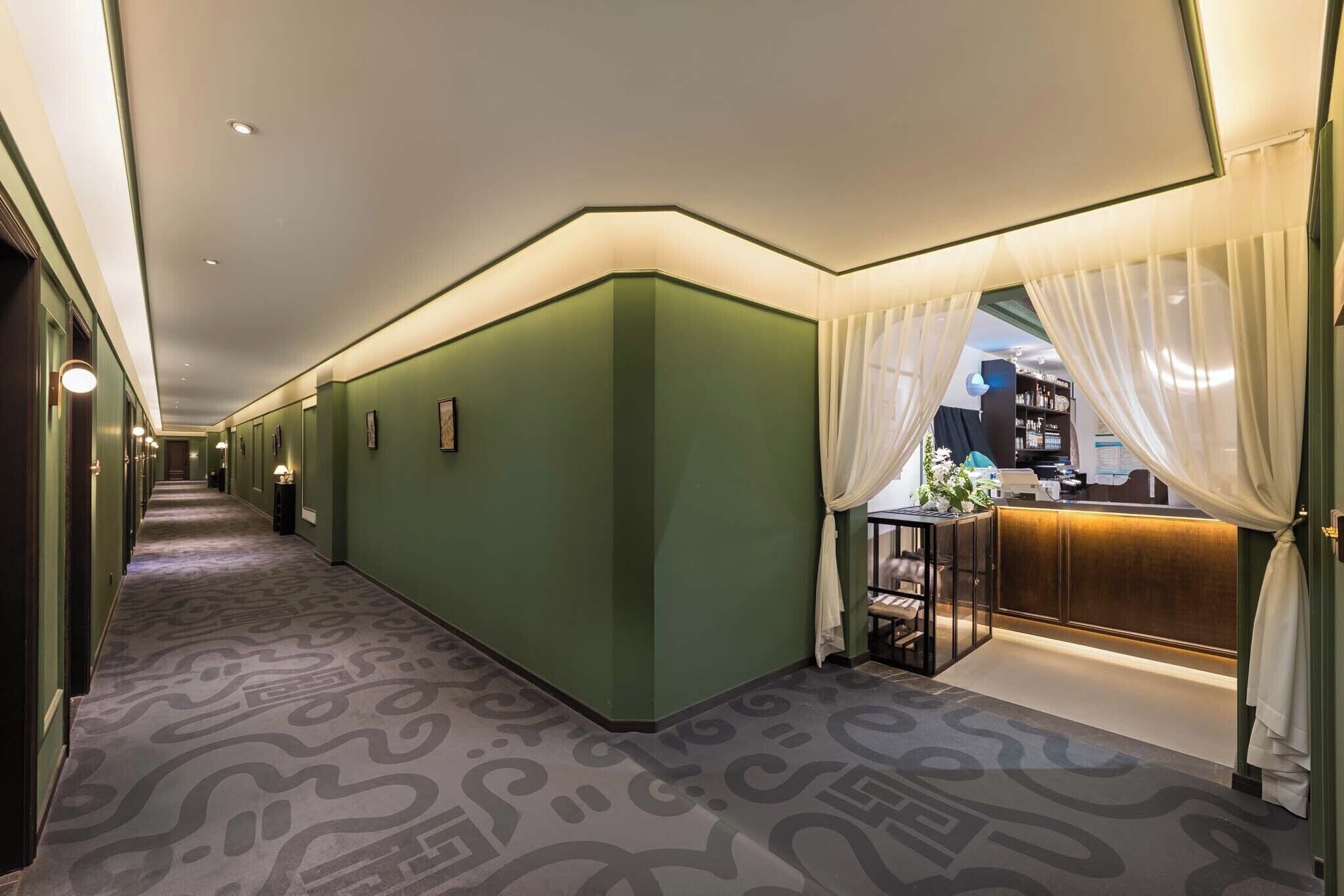 A hotel corridor with green walls, patterned carpet, a reception desk, and a visible bookshelf.