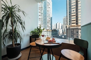 Un balcó amb una planta en test, una taula amb postre i beguda, i vista de blocs d'edificis alts.