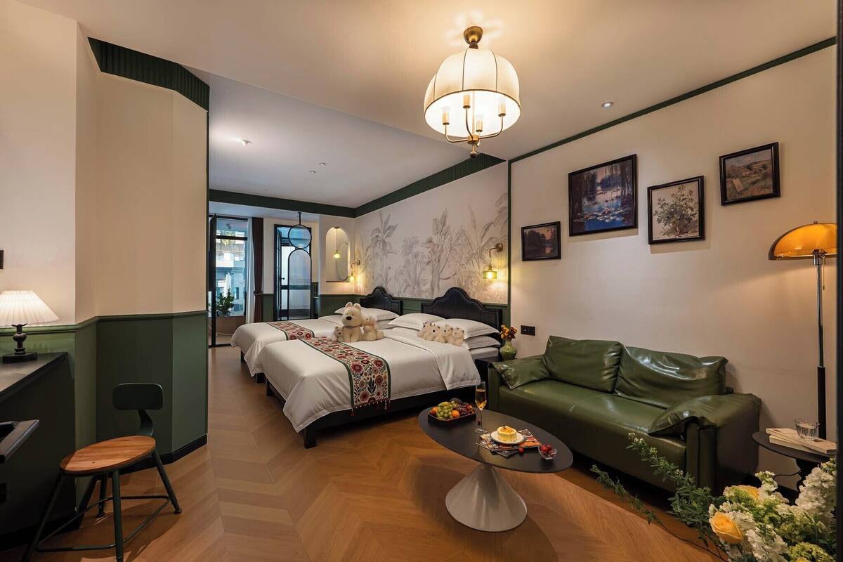 A hotel room with a bed, a green leather sofa, a dining table with a fruit bowl and a glass of wine, and a wall with framed pictures.