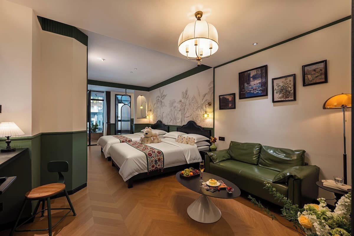 A hotel room with a bed, a green leather sofa, a dining table with a fruit bowl and a glass of wine, and a wall with framed pictures.