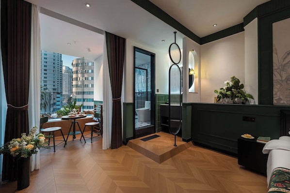 A modern hotel room with a wooden herringbone floor, a balcony with a view of high-rise buildings, a dining area with a table and chairs, and a green accent wall.