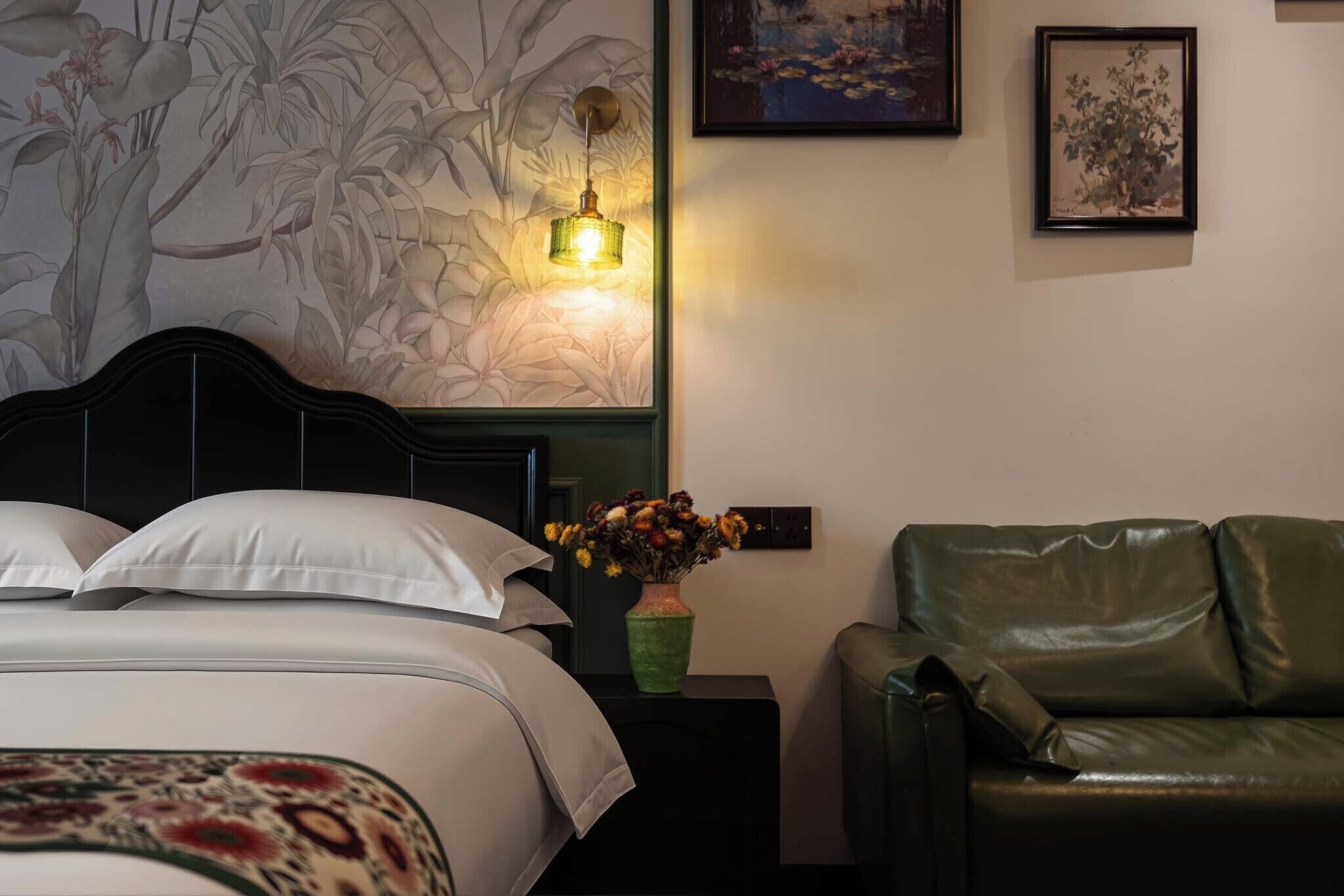 Un dormitorio con una cama, una mesita de noche con un florero, un sillón de cuero verde y decoración en la pared.
