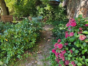 Camí de jardí amb vegetació i flors, un mur de maó i una estructura de fusta.
