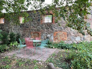 Edifici de pedra amb una finestra de rajola, seients exteriors amb caires de metall i una zona de jardí.