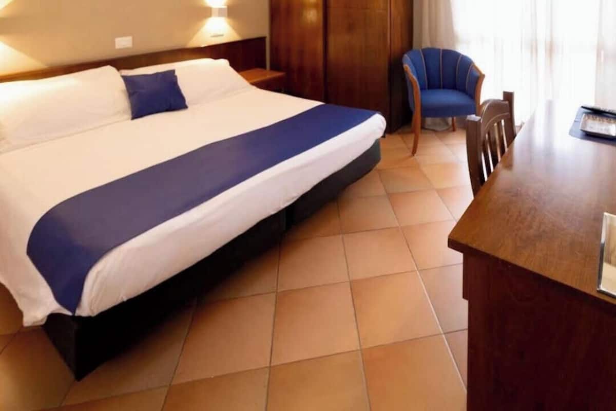Quarto de hotel com uma cama grande, uma mesa de cabeceira de madeira, uma cadeira e uma escrivaninha.