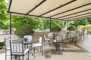En udendørs terrasse med borde og stole, omgivet af grøn vegetation og et overdækket område.