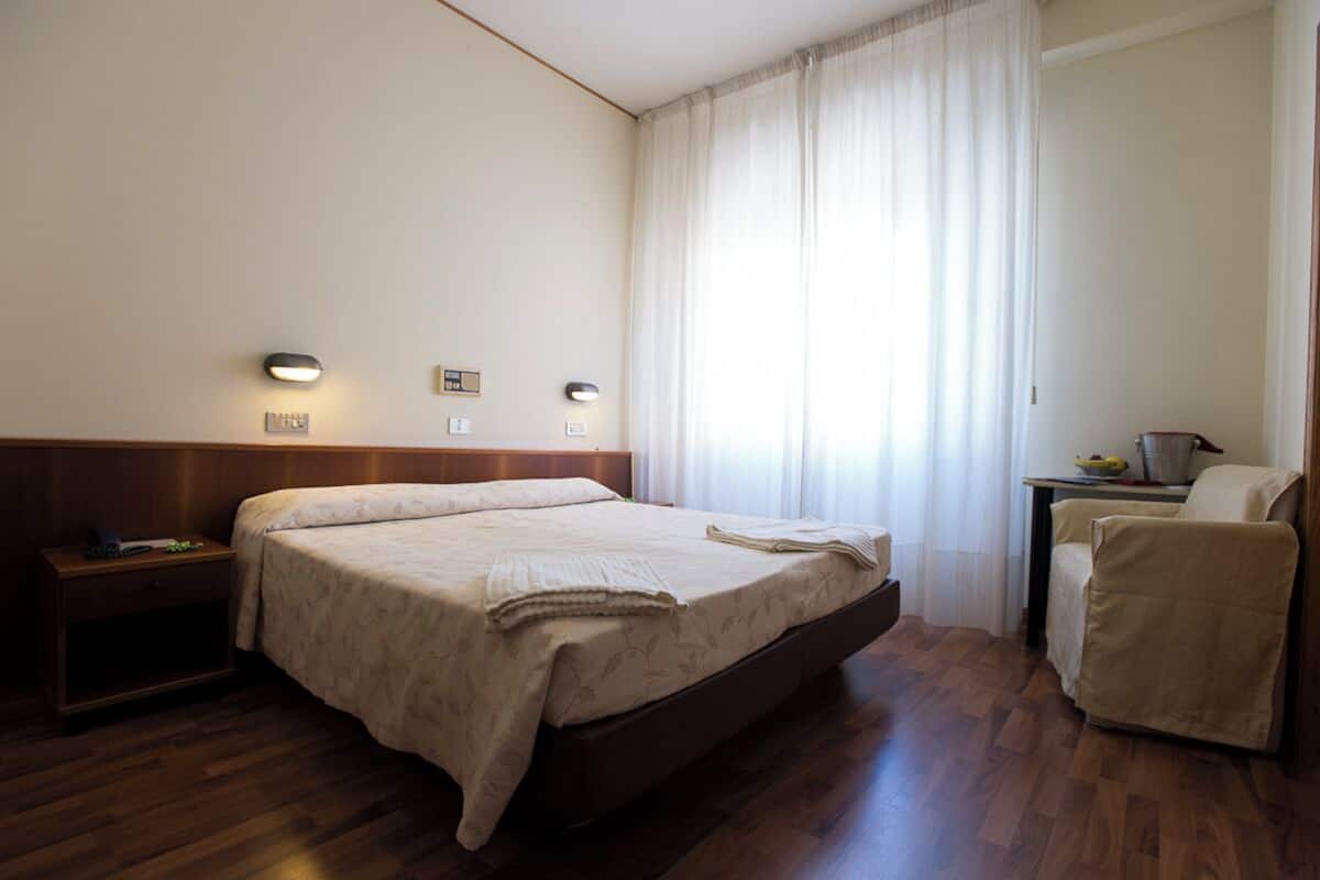 Quarto de hotel com cama, mesa de cabeceira, abajur, cadeira e janela com cortinas.