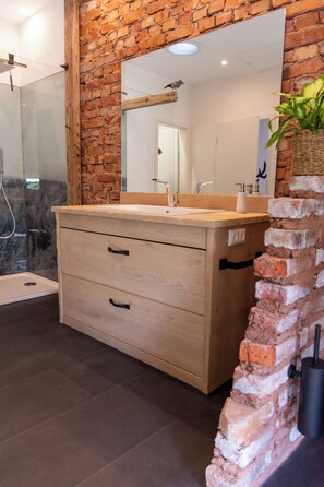 Un baño con tocador de madera, cabina de ducha con mampara de vidrio y pared decorativa de ladrillo.