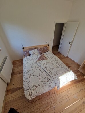 Un dormitorio con piso de madera, una cama con ropa de cama estampada, un radiador y una puerta que da a otra habitación.