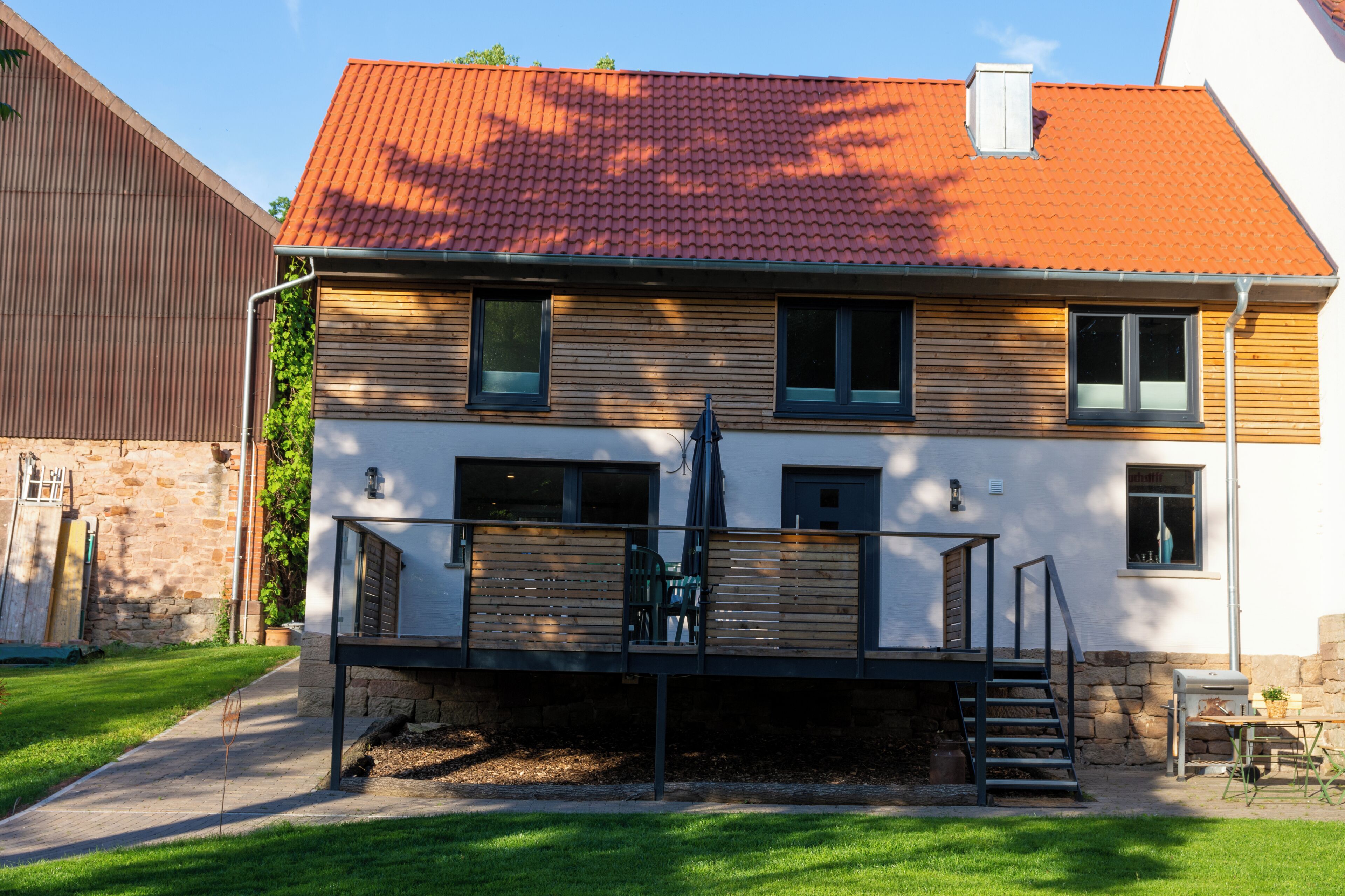 Ein zweistöckiges Haus mit rotem Ziegeldach, einer Holzterrasse und einer Steinmauer.