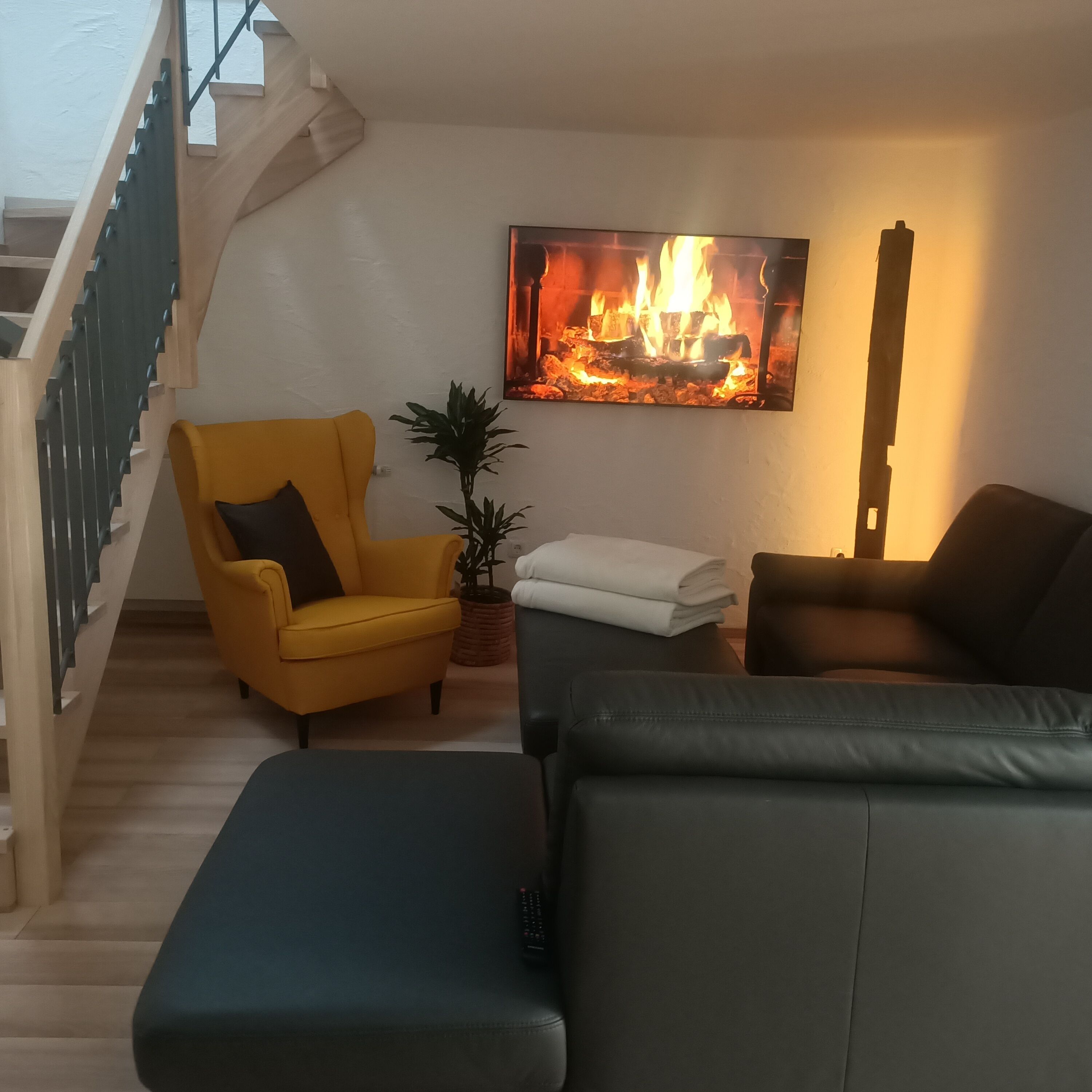 Ein Wohnzimmer mit Treppe, einem gelben Sessel, einer schwarzen Couch, einer Pflanze und einem Kamin.