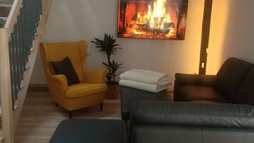 Ein Wohnzimmer mit Treppe, einem gelben Sessel, einer schwarzen Couch, einer Pflanze und einem Kamin.