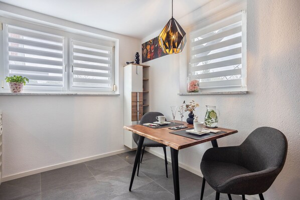A dining area with a wooden table, two chairs, a pendant light, and a window with blinds.