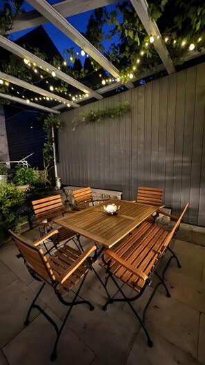 An outdoor patio with a wooden table and chairs, string lights, and a wooden fence.