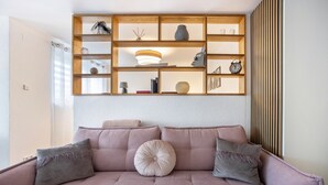 A living room with a sofa, cushions, a wooden shelf with decorative items, and a wall with vertical blinds. - Apartment 'Wohlfühlappartement Sonnenhalde' with Private Terrace, Garden & Wi-Fi (Sigmarszell)