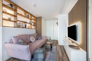 Moderni olohuone, jossa on vaaleanpunainen sohva, puuhyllyt, litteänäyttöinen televisio ja pieni pyöreä pöytä, jonka päällä on viinipullo.