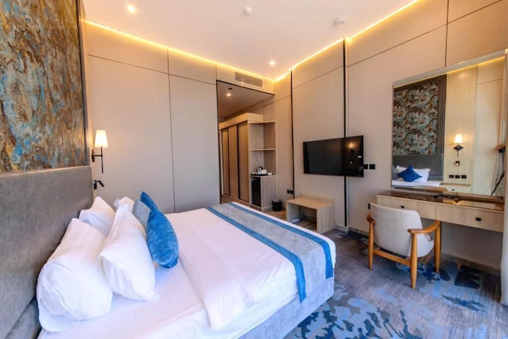 A modern hotel room with a bed, a desk, a chair, a TV, and a large mirror.