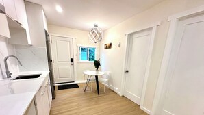 A kitchen with white cabinets, a sink, and a window. There is a dining table with chairs and a potted plant. - Cozy new 2-bedroom suite with AC & big private yard in charming Burnaby (Burnaby)