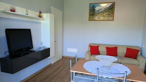 Ein Wohnzimmer mit Fernseher, einer Couch mit roten Kissen, einem Gemälde an der Wand und einem Tisch mit weißer Spitzenleinentuch.
