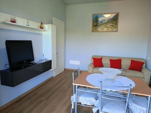 Ein Wohnzimmer mit Fernseher, einer Couch mit roten Kissen, einem Gemälde an der Wand und einem Tisch mit weißer Spitzenleinentuch.