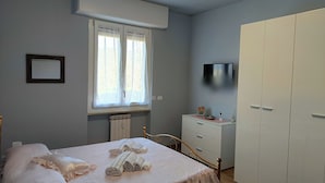 Ein Schlafzimmer mit Bett, Kommode, Schrank, einem Fenster mit Vorhängen, einem Heizkörper und einem an der Wand befestigten Fernseher.