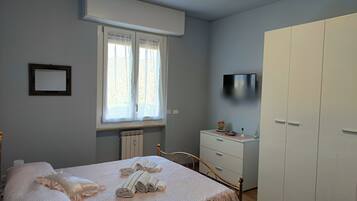 Una camera da letto con un letto, una cassettiera, un armadio, una finestra con tende, un termosifone e una televisione fissata al muro.