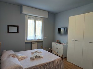 Ein Schlafzimmer mit Bett, Kommode, Schrank, einem Fenster mit Vorhängen, einem Heizkörper und einem an der Wand befestigten Fernseher.