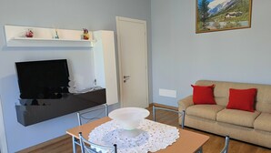 Ein Wohnzimmer mit Fernseher, einer Couch mit roten Kissen, einem Tisch mit weißer Spitze und einem Bild an der Wand.
