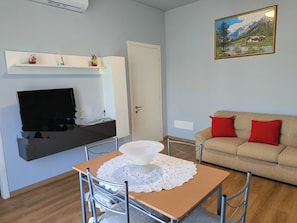 Ein Wohnzimmer mit Fernseher, einer Couch mit roten Kissen, einem Tisch mit weißer Spitze und einem Bild an der Wand.