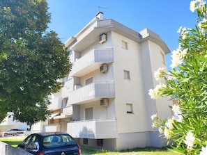Ein mehrgeschossiges Wohngebäude mit Balkonen, ein Auto davor und ein Baum mit weißen Blüten.