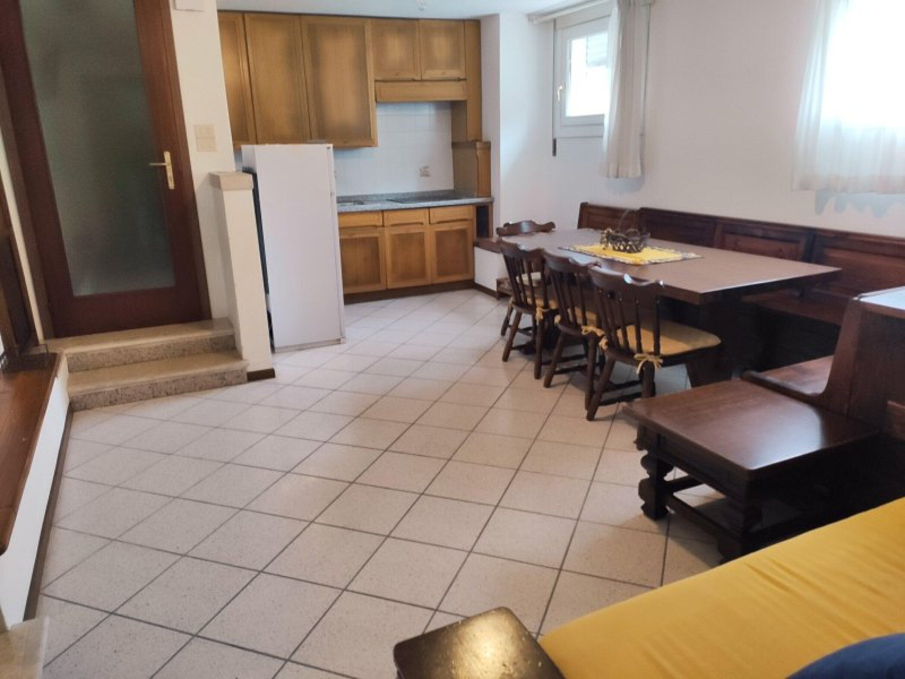Une cuisine de taille réduite, dotée de meubles en bois, d’un réfrigérateur, d’une table à manger avec des chaises et d’une fenêtre avec des rideaux.