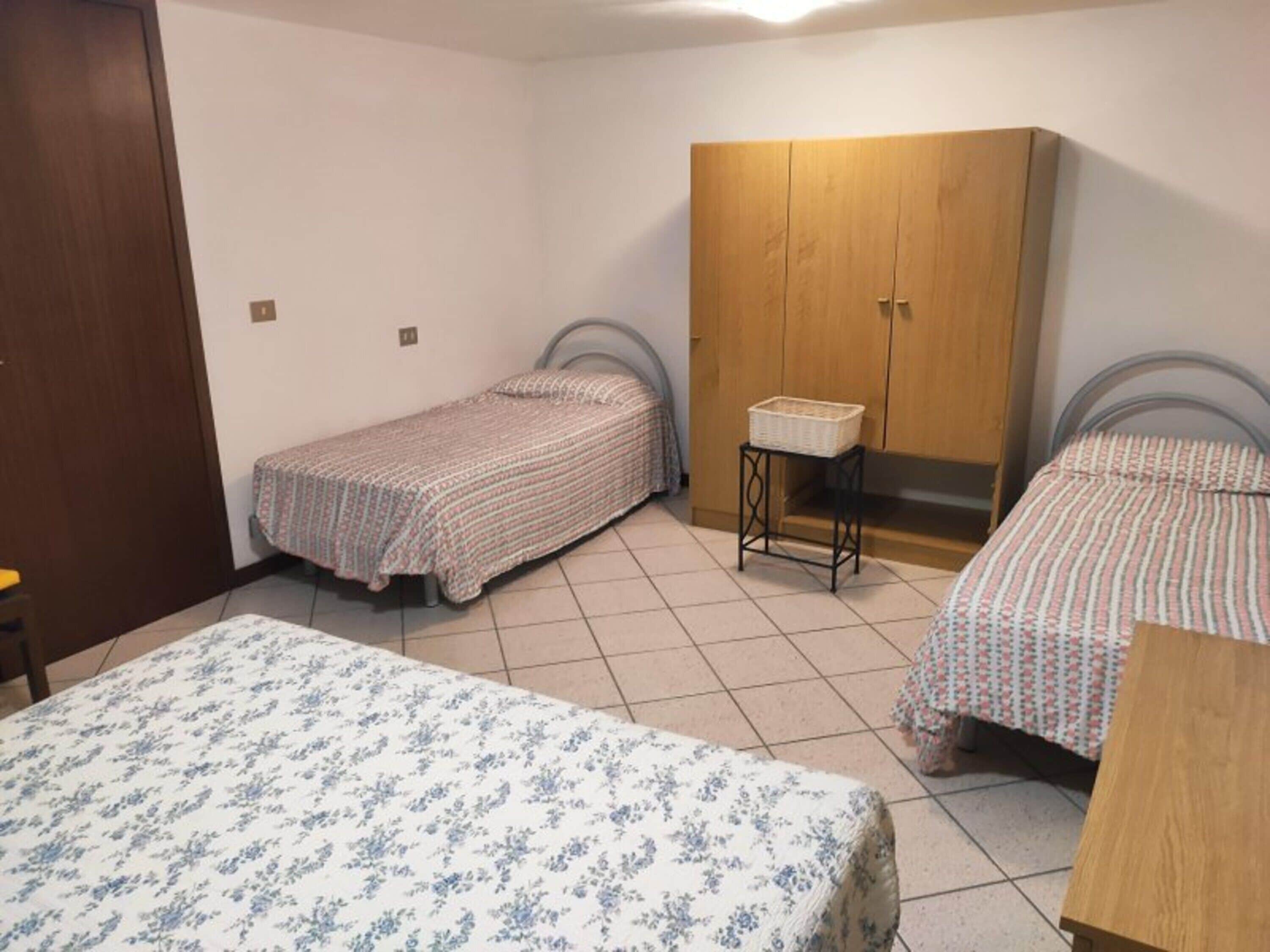 Une chambre avec deux lits simples, une armoire en bois, une petite table avec un panier, et un sol carrelé.