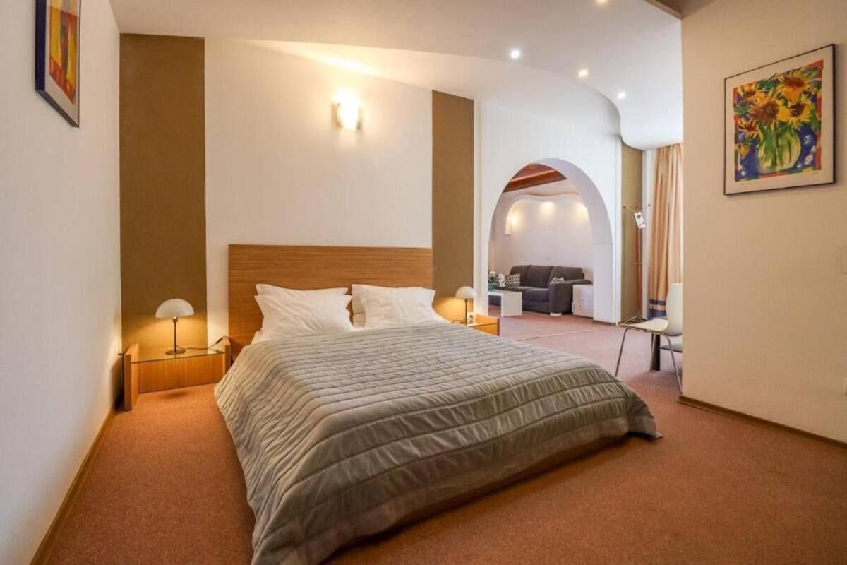 A hotel room with a large bed, a desk, and a chair. There is a painting on the wall and a lamp on the desk.