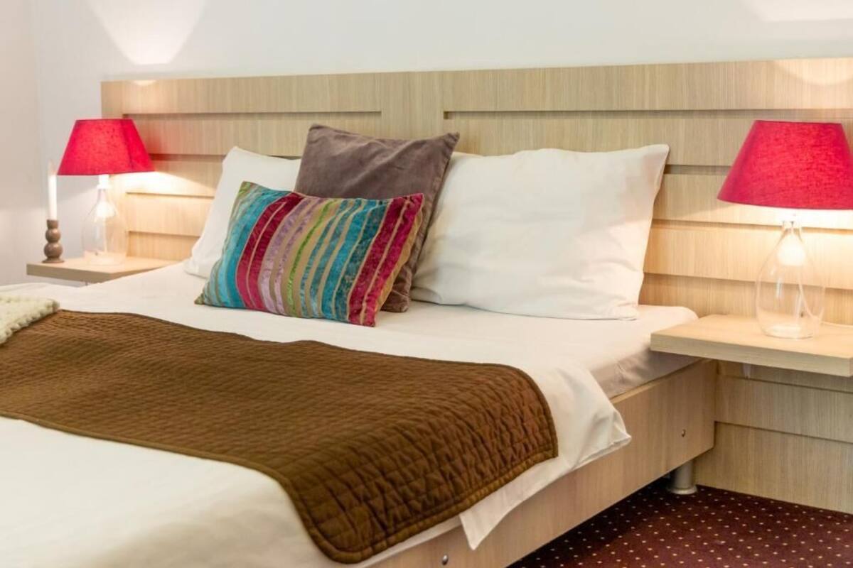 A neatly made bed with a brown blanket, a colorful striped pillow, and a white pillow. There are two bedside tables with lamps, one red and one clear glass.