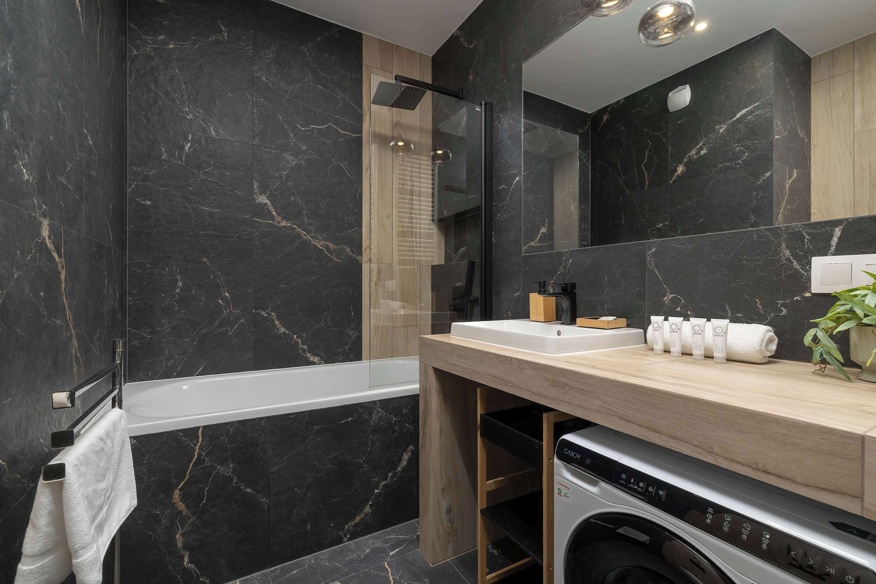 A modern bathroom with a black marble wall, a wooden vanity, a large mirror, a bathtub, and a washing machine.