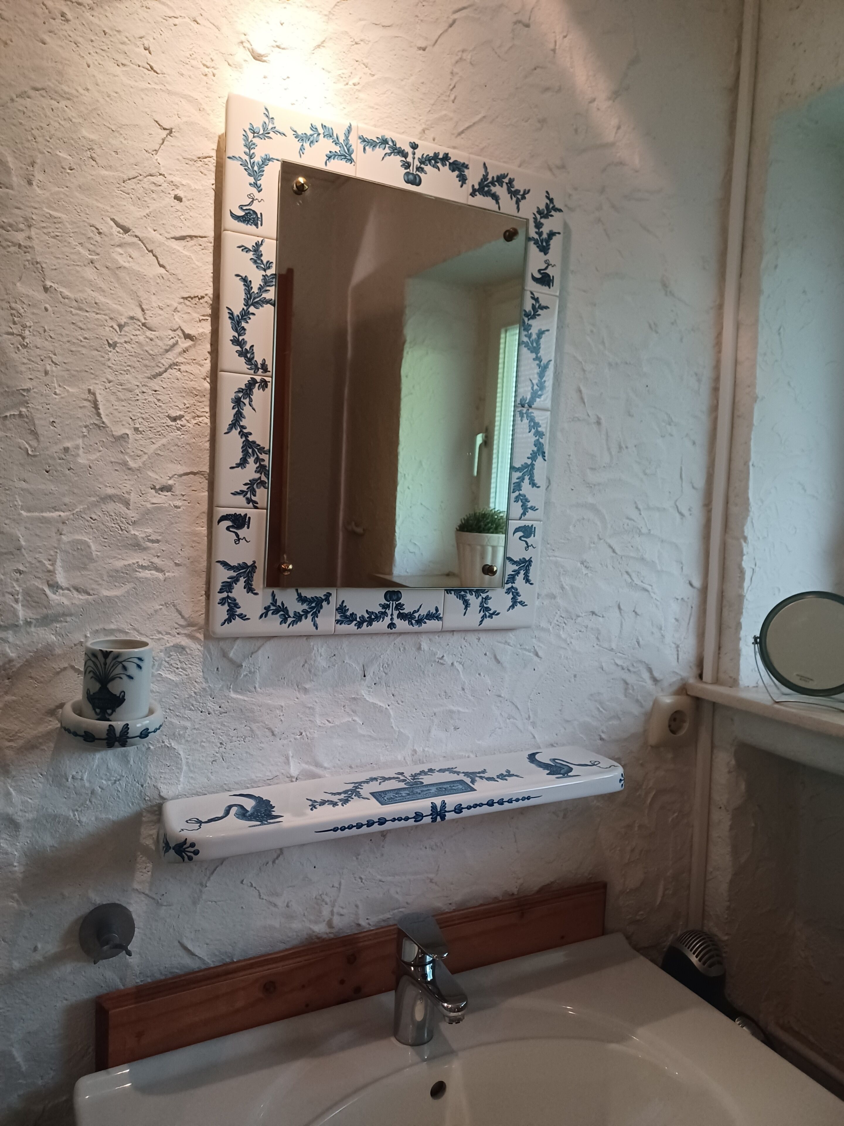Un baño con pared blanca texturizada, un espejo con marco floral azul, un lavamanos blanco con encimera de madera y una pequeña planta en maceta.