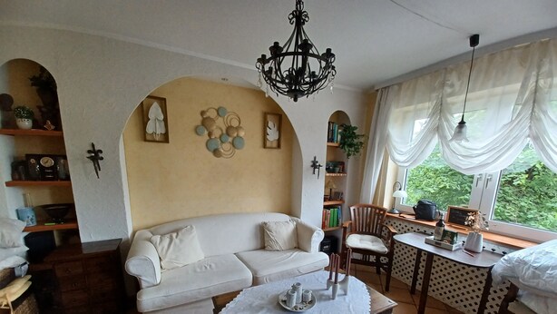Ein gemütliches Wohnzimmer mit einer Couch, einem Holztisch mit Spitzenunterlage, einer Chandelierleuchte und Blick durch ein Fenster mit transparenten Vorhängen.