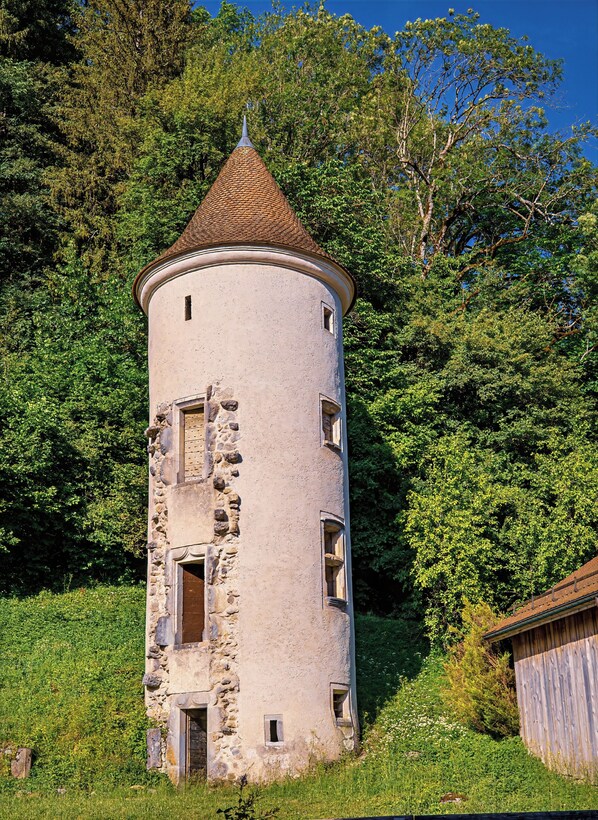A historic tower with a conical roof, multiple windows, and a stone base, surrounded by lush greenery.