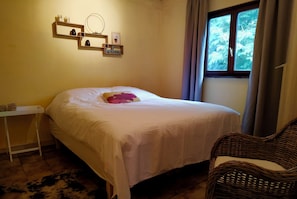 A bedroom with a bed, a bedside table, a wicker chair, a window with curtains, and wall-mounted shelves with items.