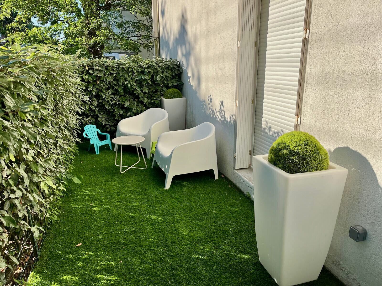 Eine kleine Terrasse mit künstlichem Rasen, weißer Gartenmöbel und einem Blumentopf.