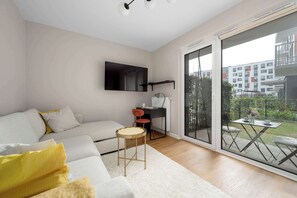 Una sala de estar moderna con un sofá, una mesita redonda, un escritorio con dos sillas y un amplio ventanal corredero que da a un espacio exterior.