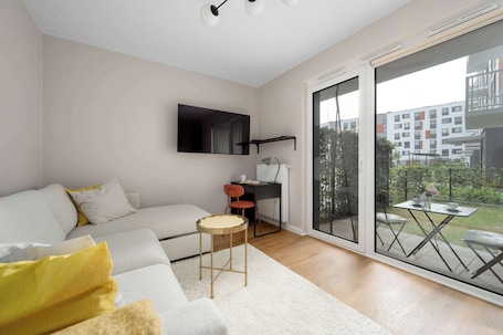A modern living room with a sofa, a small round table, a desk with two chairs, and a large sliding glass door leading to an outdoor area.. Cozy Studio with a Garden | A Place to Work
